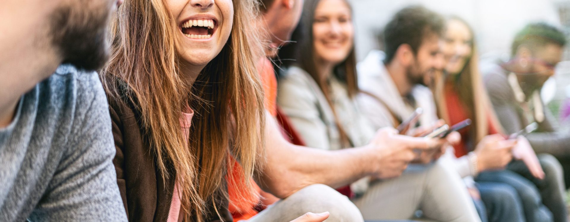 Group of trendy young people chatting together sitting on a bench outdoors. Students having fun together. Focus on a blonde girl smiling with open mouth