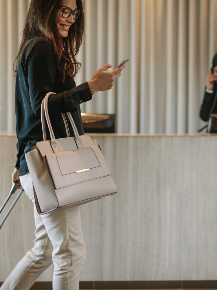Woman using mobile phone and pulling her suitcase in a hotel lobby. Female business traveler walking in hotel hallway.